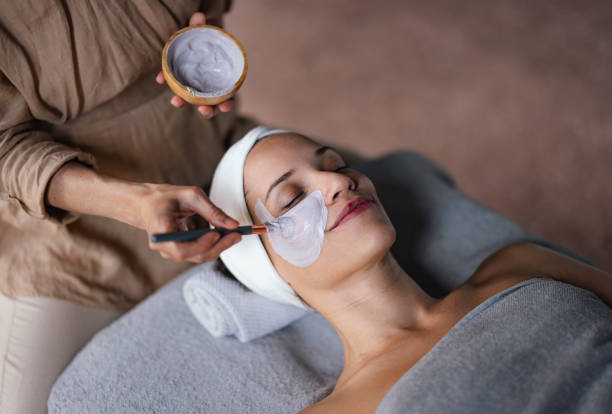 Beautiful Woman Enjoying Receiving a Facial Treatment at the Spa Close up shot of a beautiful smiling woman lying with her eyes closed while another woman is applying a face mask on her in the spa center. She is relaxed and enjoys the treatment. blank mask stock pictures, royalty-free photos & images
