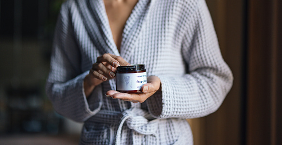 Close up shot of an unrecognizable woman standing in a bathrobe and holding a jar of face cream.
