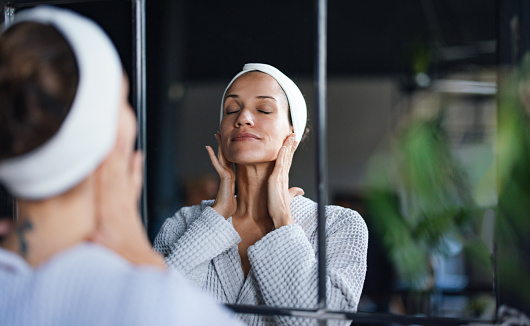Beautiful woman standing in front of the mirror at the spa with her eyes closed, touching her skin after a face mask treatment.
