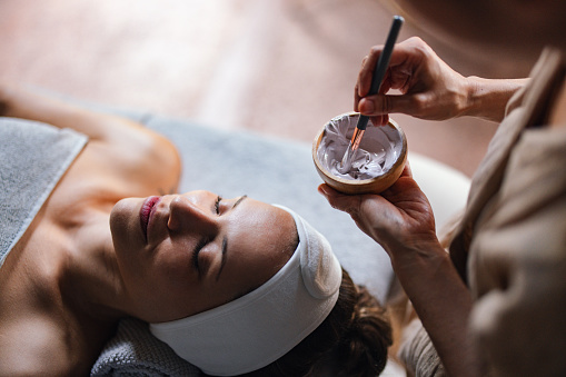 Close up shot of a beautiful woman lying with her eyes closed while another woman is applying a face mask on her in the spa center. She is relaxed and enjoys the treatment.a