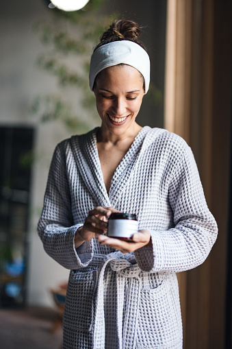 Portrait of a beautiful cheerful woman standing in a bathrobe at the spa and holding a jar of face cream. She is looking down and smiling.