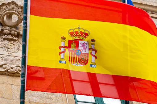 Spanish flag waving in front of a stone building. Coat of arms on flag and carved on facade. Noia Town hall, A Coruña province, Galicia, Spain.