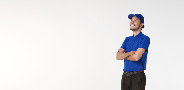 Smiling asian delivery man in blue uniform standing with arms crossed isolated on white background. looking up copy space.