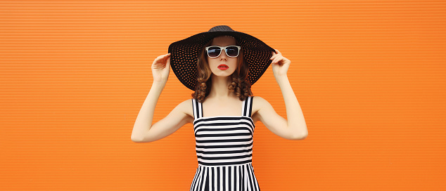 Portrait of beautiful young woman posing wearing black summer straw hat and dress on orange background