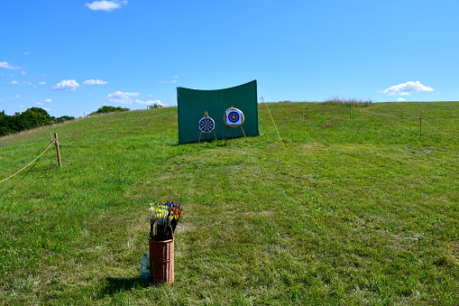 A close up on a shooting range located in the middle of a vast field or meadow next to some hay bales, with targets, bows, arrows, flags and other equipment  visible on a sunny summer day