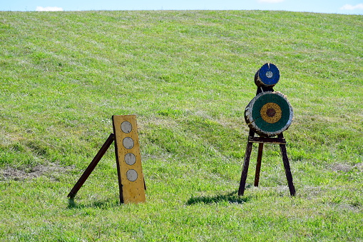 A close up on a shooting range located in the middle of a vast field or meadow next to some hay bales, with targets, bows, arrows, flags and other equipment  visible on a sunny summer day