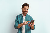 Smiling man in casual clothes use his mobile phone while standing on white background