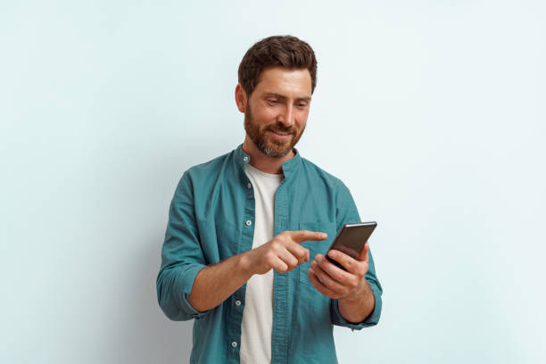 homem sorridente em roupas casuais usar seu telefone celular enquanto está em pé no fundo branco - men one person surprise looking - fotografias e filmes do acervo