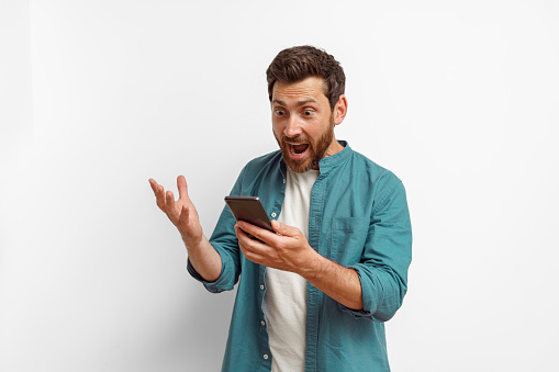 Man showing surprised gesture while looking on mobile phone on white background. High quality photo