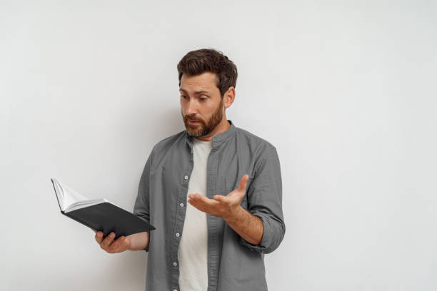 istock Baffled man in casual clothes standing with open notepad and looking on it on white background 1590551461