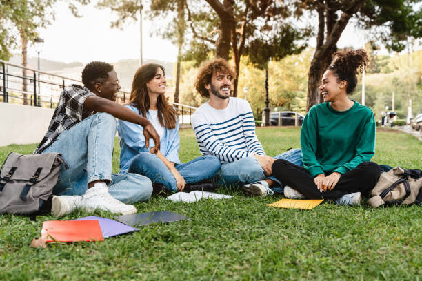Group of diverse students sitting on grass at a campus park, talking and smiling with notebooks and backpacks.