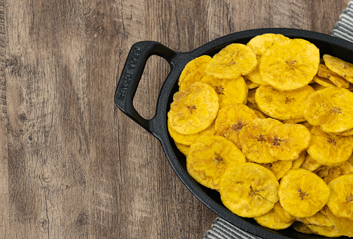 Fried banana chips in a black plate. Crispy Banana Chips. Fried sliced banana. Top view of typical traditional dish of Latin American gastronomy called Chifles or fried tostones on a wooden table.