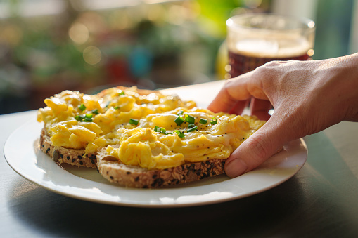 A cropped image of a woman's hand holding a piece of toasted bread with scrambled eggs on top, as she enjoys a healthy breakfast in the morning