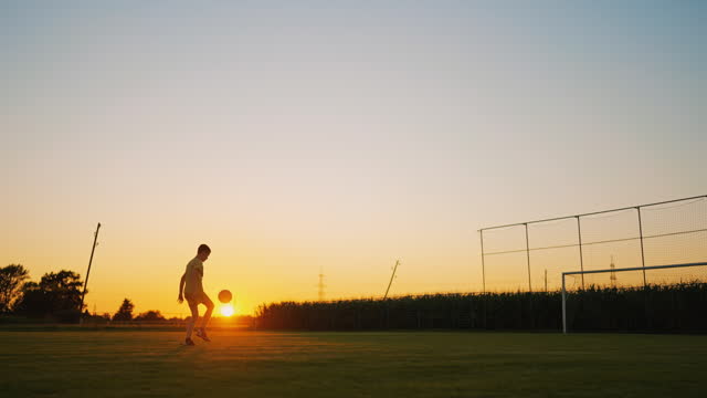 Young Boy with Soccer Ball Practicing Juggling and Heading on Sports Ground at Sunset