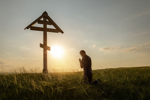 Christian man praying in the meadow at sunset. Man of prayer outdoor. Prayer at the Cross.