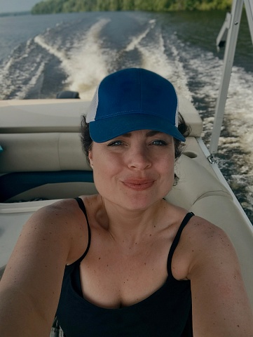 Selfie point of view of a cute middle aged woman wearing a baseball cap and riding in a moving pontoon on The Menominee River on the border of Upper Michigan and Wisconsin.
