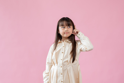 An adorable little Asian girl in a cute dress is standing against an isolated pink background with a cute thoughtful face.