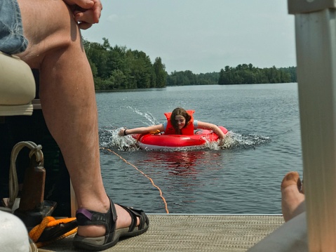 Teen girl, on a swim float, tied with a tow rope to a pontoon boat(deck of the boat point of view with other passengers feet seen on the deck)on The Menominee River on the border of Northern Michigan and Wisconsin. The girl is paddling herself in toward the boat.