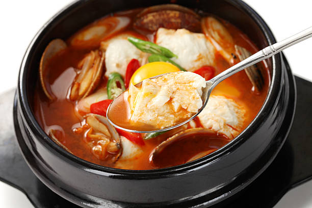 A close-up, high-angle shot of a small white bowl of soup with a clear broth and chopped vegetables. The bowl is set on a wooden table next to a blurry, out-of-focus Bibimbap bowl, and a drink, highlighting a simple yet essential part of a traditional Korean meal.