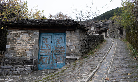 Traditional old stone building with aged peeled wooden blue door and stone roof next to paved street Epirus travel Greece, nature, winter day.