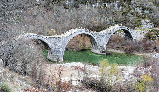 Kalogeriko or Plakidas ancient stone bridge with three arch over Voidomatis river water, Zagoria Epirus Greece. Dry tree background.