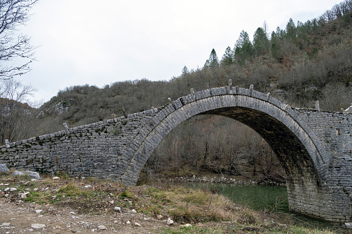 Kalogeriko or Plakidas ancient stone bridge. View of one of three arch over Voidomatis river water, Zagoria Epirus Greece. Dry tree background.