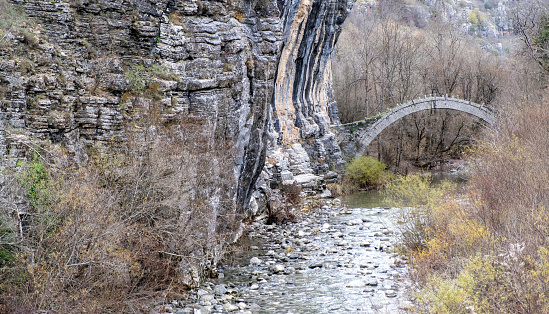 Greece, Lazaridi Kontodimou medieval arched stone bridge over river at Vikos Canyon Epirus. Rocky winter landscape, dry tree background.