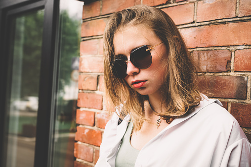 Portrait of a young woman in a white shirt and sunglasses on the background of the exterior of the brick wall of the building.