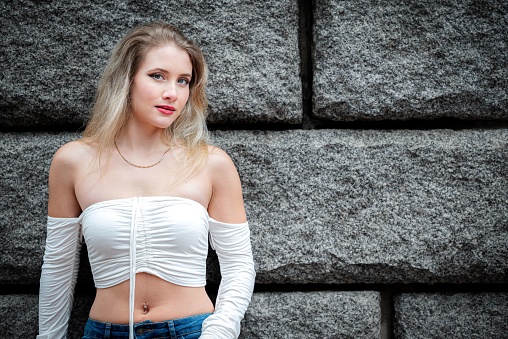 A young female with blonde hair poses in a casual outfit consisting of a white top and blue jeans