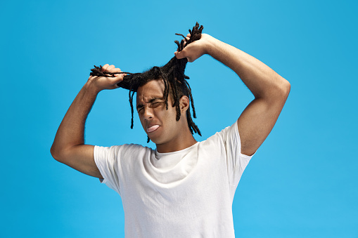 Meme look. Young guy pulling hair, dreads and sticking out tongue against blue studio background. Grimacing face. Concept of youth, human emotions, lifestyle, fashion, facial expressions, ad
