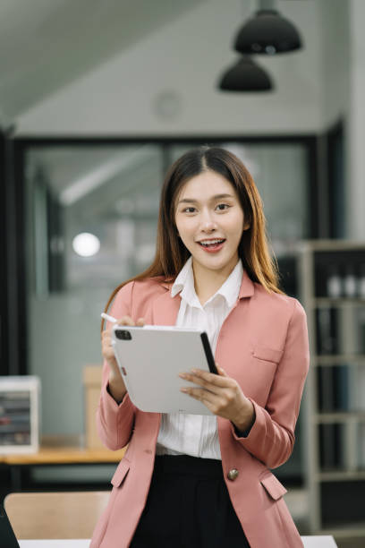 istock Asian woman holding tablet working at desk, standing smiling looking at camera. 1591522553