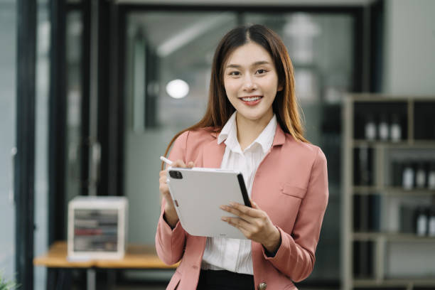 istock Asian woman holding tablet working at desk, standing smiling looking at camera. 1591522555