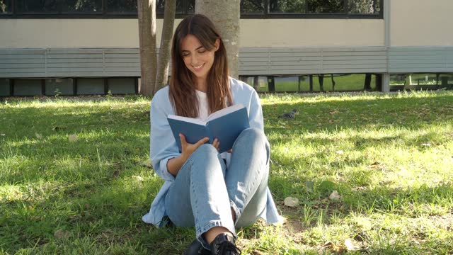 Young smiling woman reading a book in a park under a tree