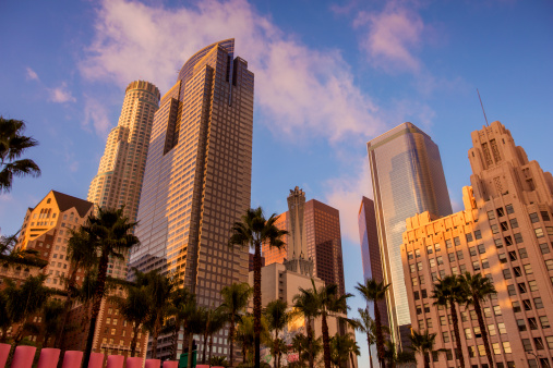 Downtown Los Angeles skyscrapers with palm trees and Pershing Square in the foreground with clouds and a blue sky at sunset. Tropical flair.