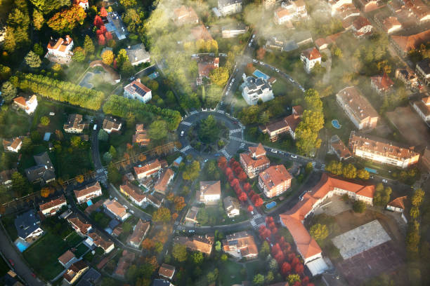 Aerial perspective from hot air balloon floating over traffic circle, shapely layout for residential buildings, autumn leaf colors, and patchy low-lying fog.