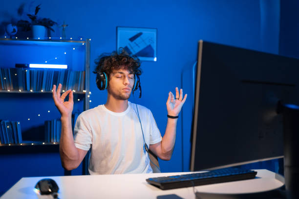 istock Arabic guy gamer meditates sitting before computer in a headset at night in neon light, closed his eyes making yoga. 1591662377