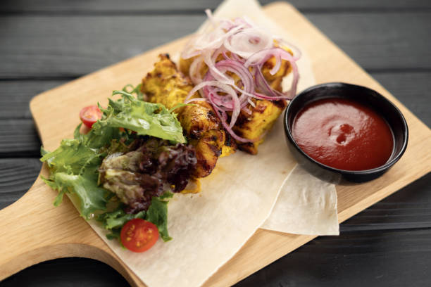 Healthy barbecued chicken kebabs served with a tortilla and fresh lettuce and tomato salad, on a dark wooden background, top view