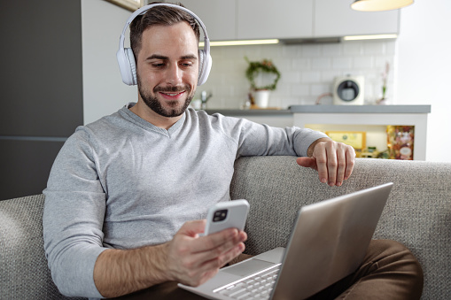 Young man relaxing at home. He is using phone and a laptop.