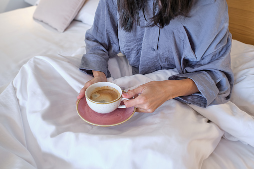 Unrecognizable awaken woman in sleepwear resting on soft white bed with coffee mug in hands and drinking coffee at bedroom at home in morning