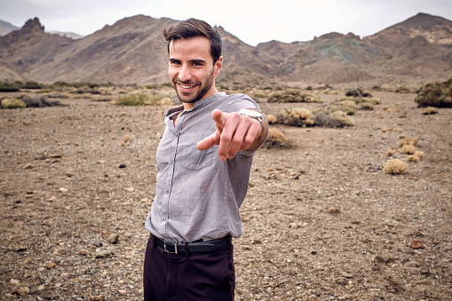 Positive smiling bearded male traveler standing in dried valley near highlands pointing with index finger and looking at camera