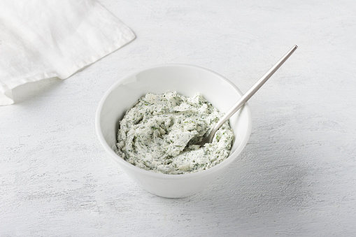 White bowl with cottage cheese cream mixed with herbs and garlic on a light gray background. Cooking appetizer or toppings for stuffing vegetables, vegetarian food.