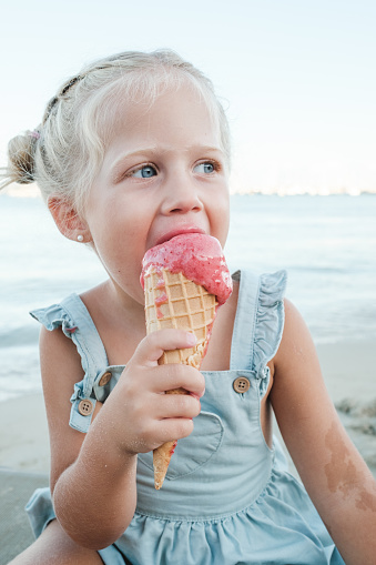 Fair haired child in blue dress biting ice cream cone while sitting on beach and looking away thoughtfully