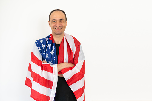 Happy relaxed satisfied handsome middle aged man standing wrapped in USA flag, celebrating national holiday, wearing urban style hoodie. Indoor studio shot isolated on white background. High quality photo