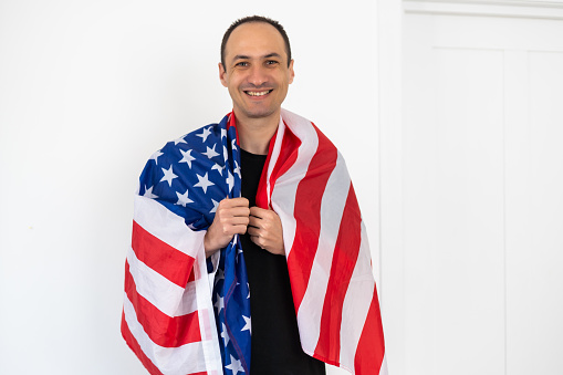 Celebrating an Independence day. Stars and Stripes. Young man with the flag of the United States of America isolated on white studio background. Looks crazy happy and proud as a patriot of his country. High quality photo