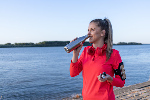 Beautiful young woman in her 20s stands on a jogging path by the river in fitness clothes and drinks water from a metal bottle. Standing elegantly on the scenic jogging path alongside the river, a beautiful young woman in her 20s, wearing fitness clothing, pauses to drink water and catch her breath
