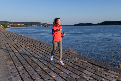 Beautiful young woman in her 20s running on a jogging path by the river in fitness clothes and holding a metal water bottle in her hand while running and training illuminated by the late afternoon sun.