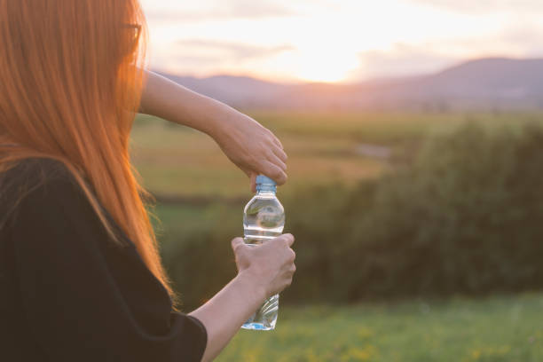 Woman is opening water bottle outdoor in nature at sunset of hot, summer day