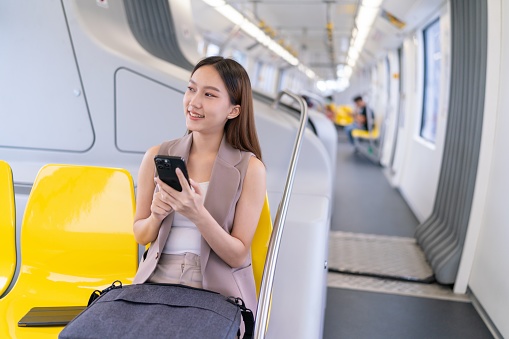 Seated in the monorail, the young Asian businesswoman's cheerful expression reveals her engagement with social media video content on her mobile phone, adding an element of excitement to her journey.
