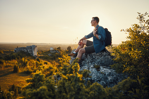 Travel and hiking concept. Adult man with backpack sitting on the edge of rock enjoying landscape during sunset