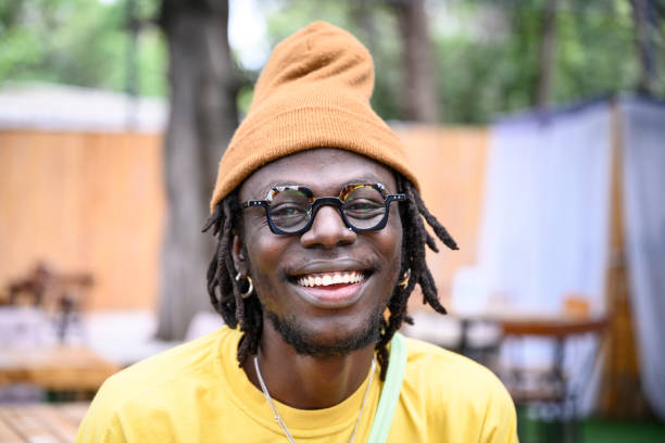 Outdoor portrait of cheerful African man in early 30s Close view of Nairobi man with medium-length locs and facial hair, wearing fashionable eyewear and knit hat, smiling at camera. kenyan man stock pictures, royalty-free photos & images